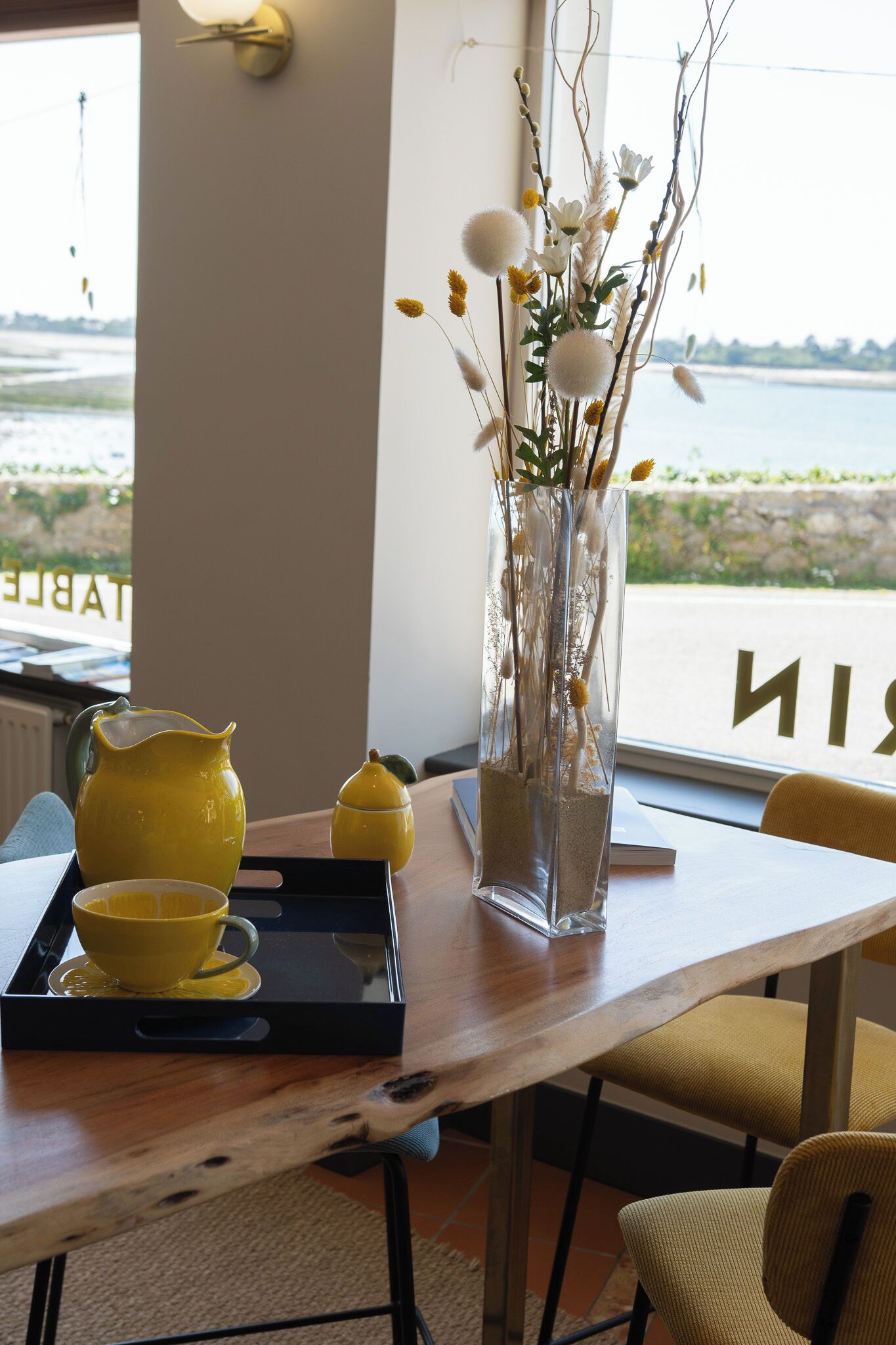 Een tafel met een gele karaf, kopje en schotel, een vaas met takken en bloemen, en een uitzicht op zee door een raam.