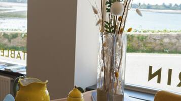 Een tafel met een gele karaf, kopje en schotel, een vaas met takken en bloemen, en een uitzicht op zee door een raam.
