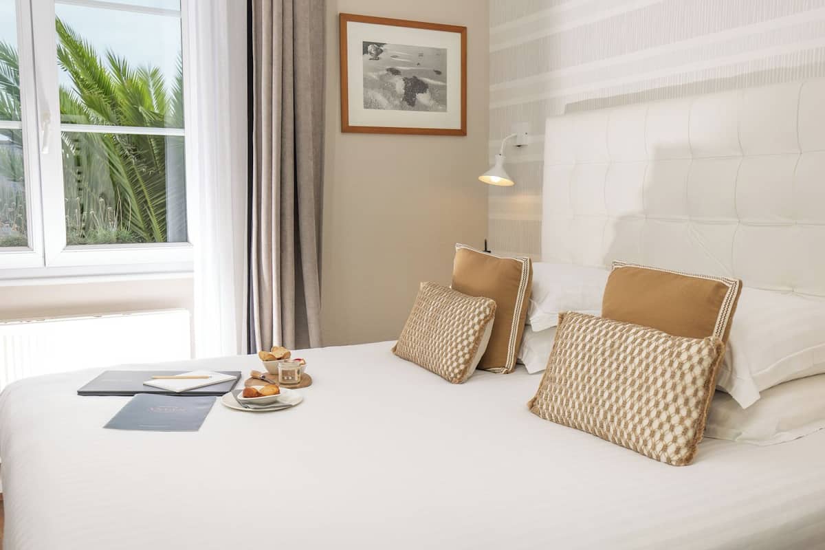 A hotel room with a large bed, a bedside table with a lamp, a framed picture on the wall, and a window with curtains.