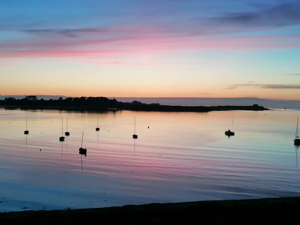 A calm body of water with several boats anchored, a shoreline with trees, and a sunset sky.