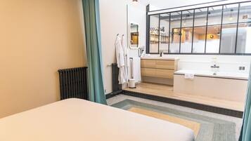 A modern hotel room with a bed, a bathroom with a bathtub, a window with a view of the interior, and a radiator.