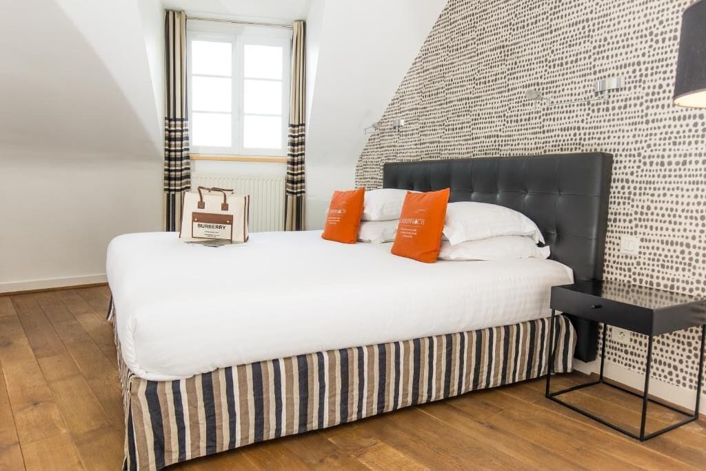 A neatly made bed with striped bedding and two orange pillows, a black headboard, and a small bedside table.