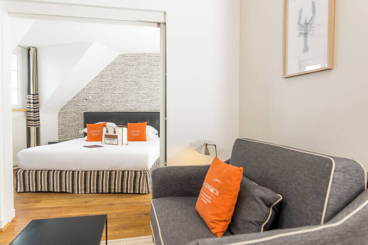 A hotel room with a bed, a grey sofa, a black table, and a staircase with a patterned wall.