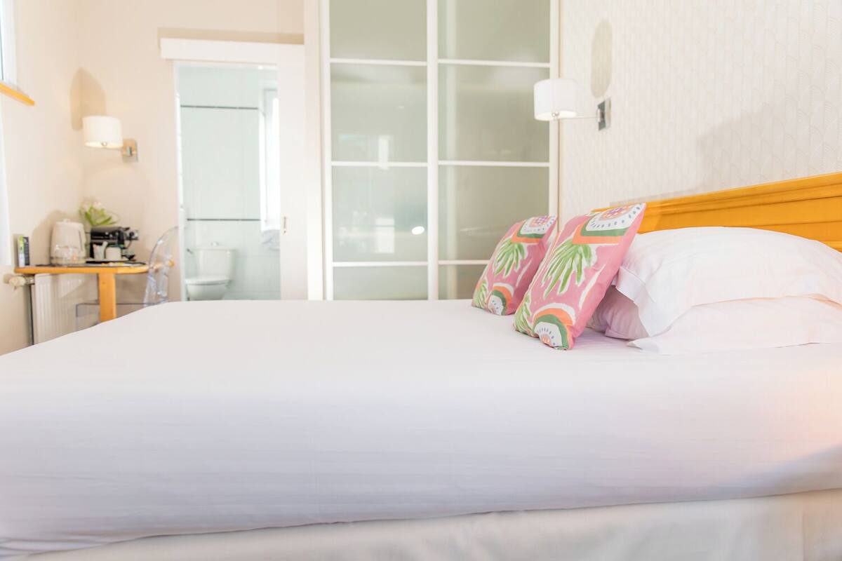 A neatly made bed with white linens and two decorative pillows, a wooden headboard, a small bedside table with a lamp, and a view of a glass door leading outside.