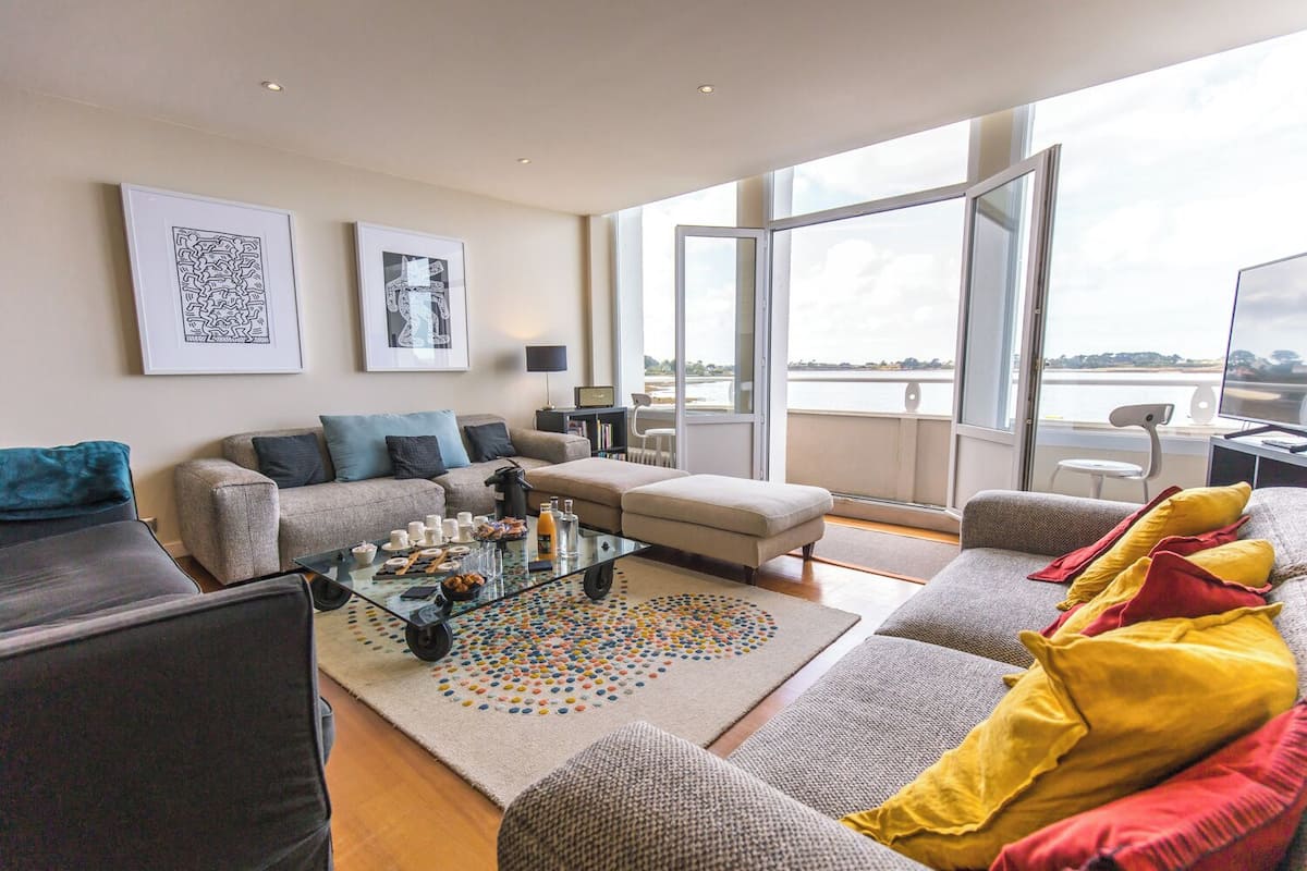 A modern living room with a sofa set, a coffee table, and a view of a body of water through large windows.