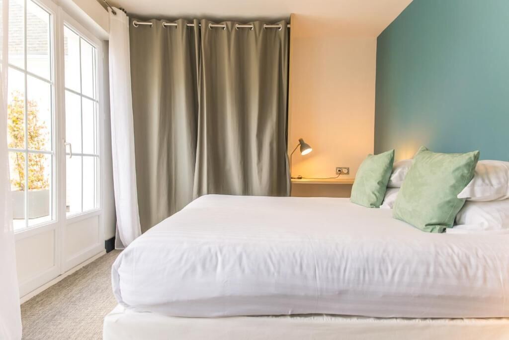 Een slaapkamer met een groot bed, een nachtkastje, een lamp en uitzicht op de natuur door een raam met gordijnen.