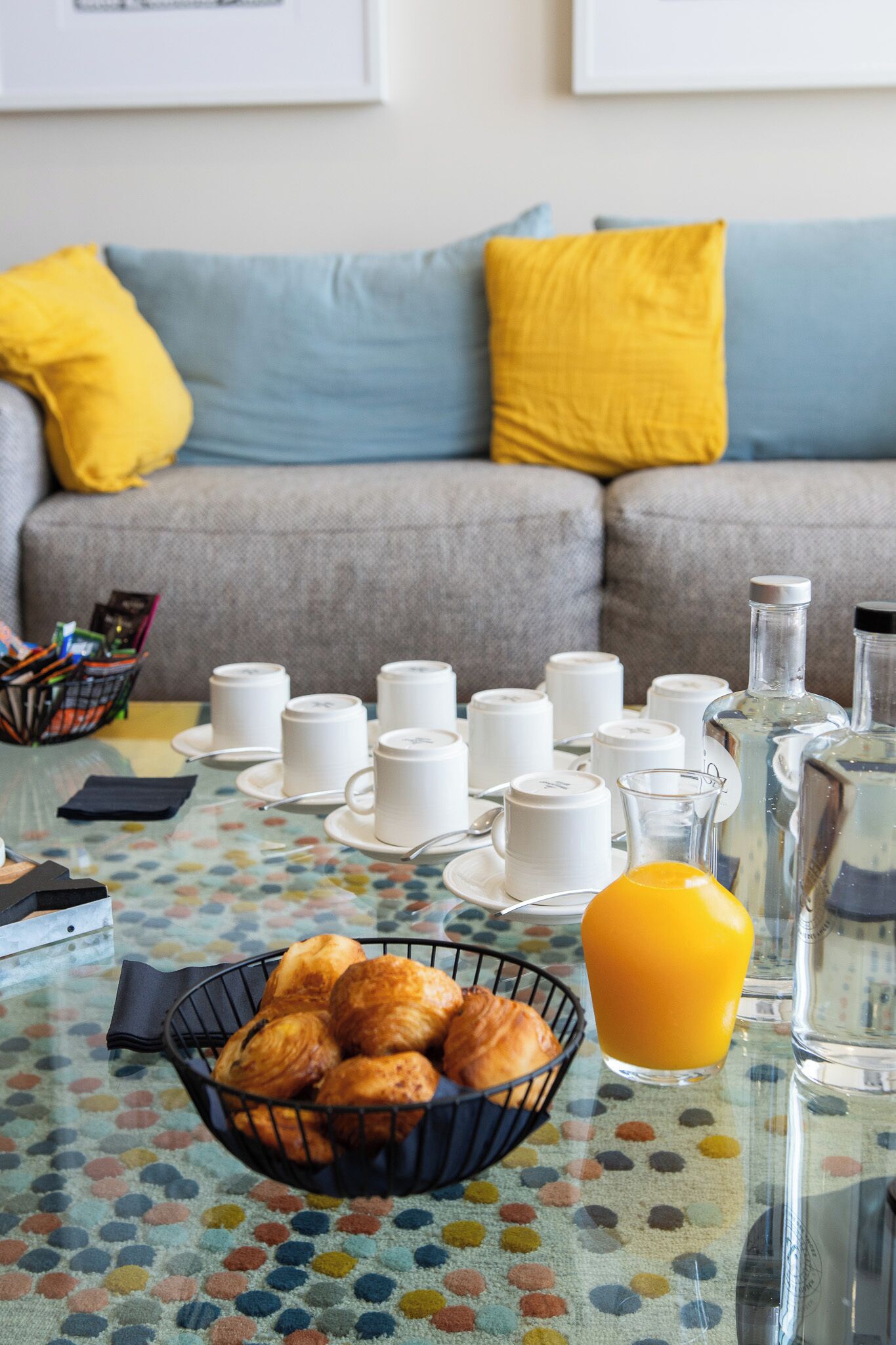 A breakfast setup with a bowl of croissants, a glass of orange juice, and multiple white cups and saucers on a table with a polka dot tablecloth.