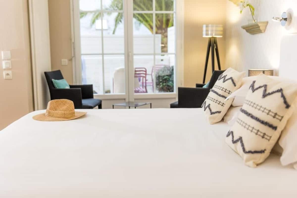 A hotel room with a bed, two chairs, a window with a view of palm trees, and a lamp on a bedside table.