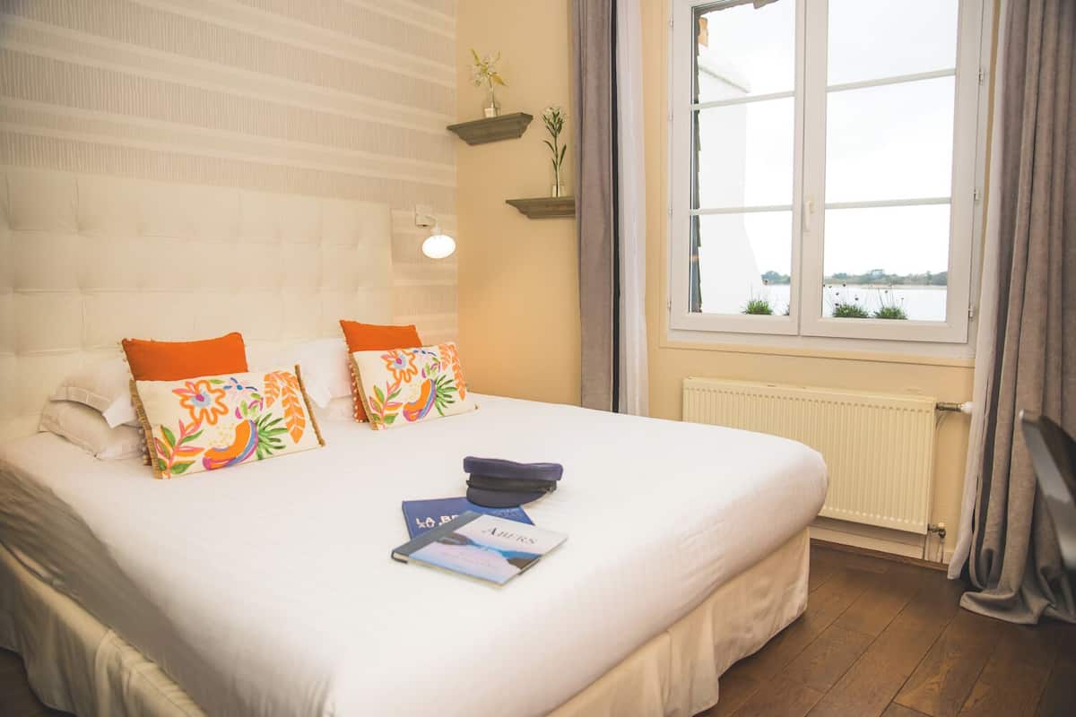 A neatly made bed with decorative pillows, a bedside table with a book and a remote, and a window with a view of a body of water.