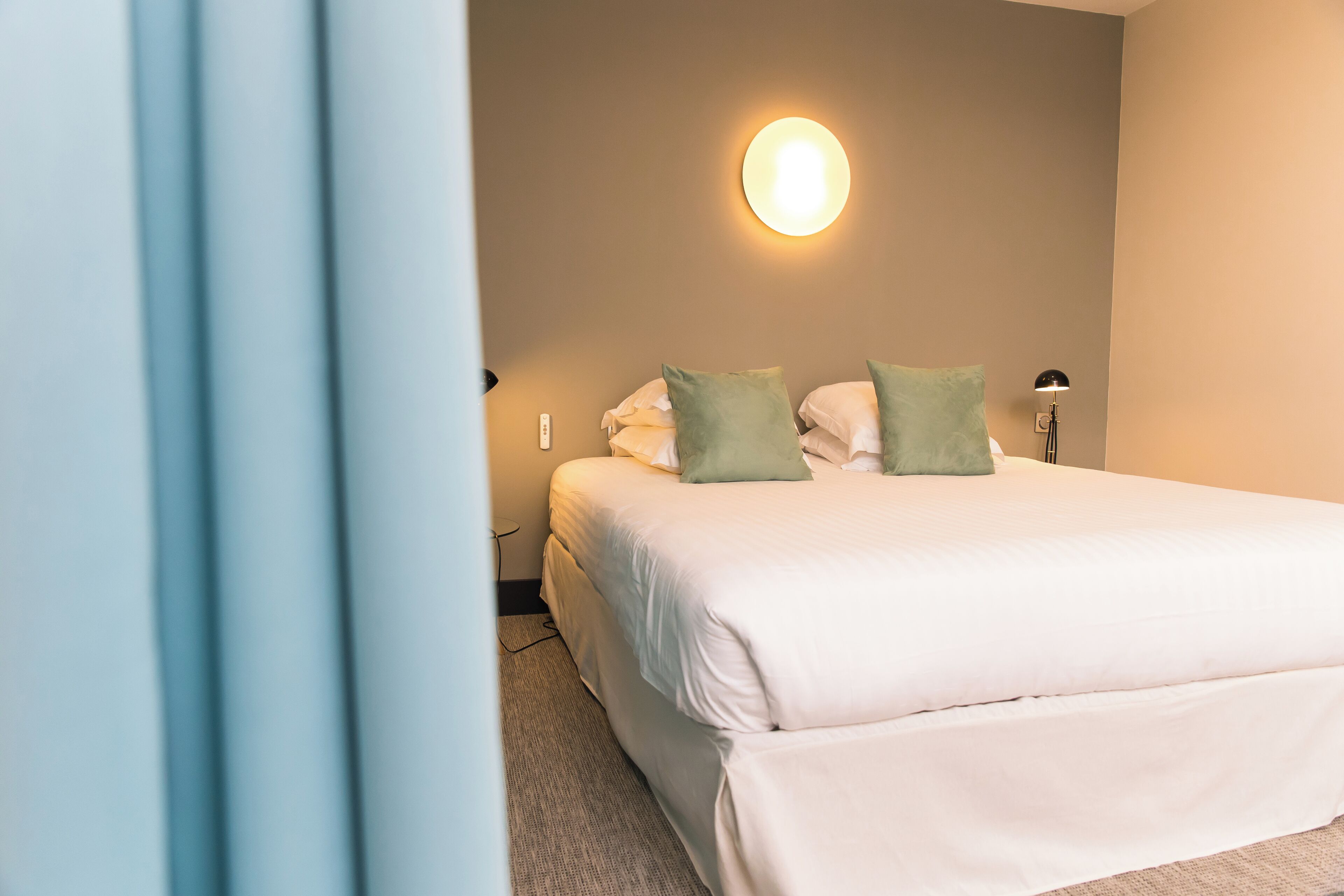 A neatly made bed with white and green pillows in a hotel room with a wall-mounted light fixture.