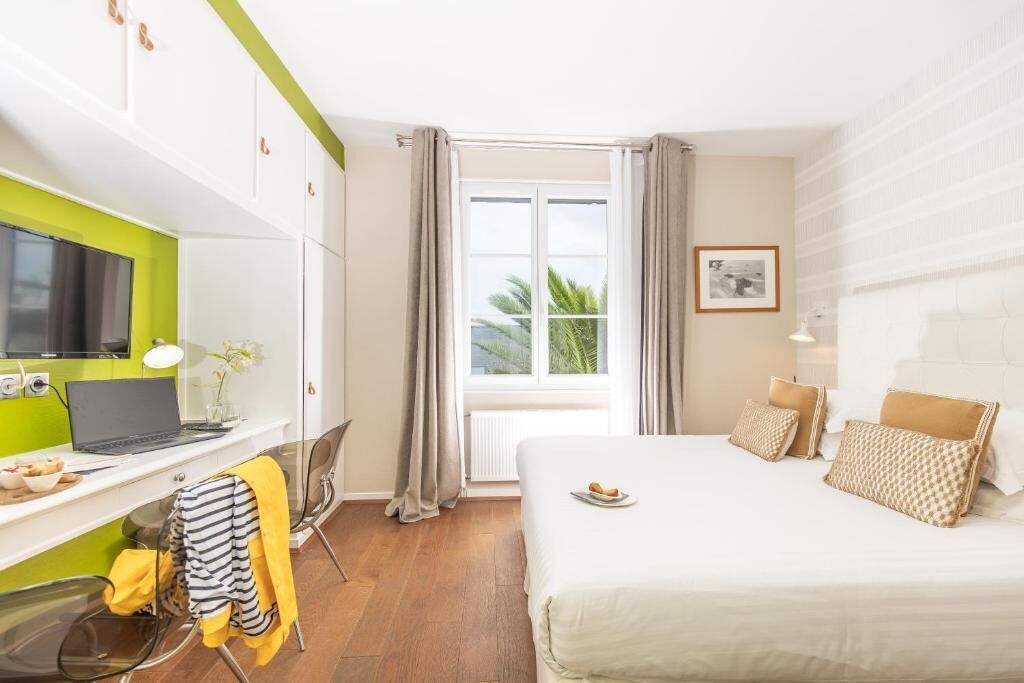 Habitación de hotel con cama, escritorio con una computadora portátil, silla, ventana con cortinas e imagen enmarcada en la pared.