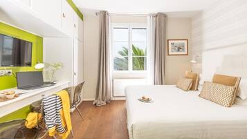 Habitación de hotel con cama, escritorio con una computadora portátil, silla, ventana con cortinas e imagen enmarcada en la pared.