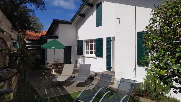 A white house with green shutters, a wooden deck with lounge chairs, and a green umbrella.