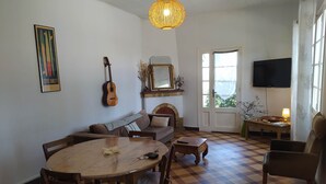 A cozy living room with a checkered floor, a round wooden dining table, a sofa, a guitar, a television, and a window with a view.