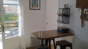A small kitchen with a table, stools, a trash can, a hanging light fixture, a framed picture, and a window with curtains.