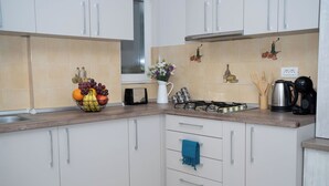 Une cuisine avec des meubles blancs, un plan de travail en bois, une cuisinière à gaz, une bouilloire et un panier de fruits.