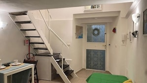 Una habitación compacta con una escalera que lleva a una puerta con paneles de vidrio decorativos. Se ve una pequeña mesa con una silla y una cama verde.