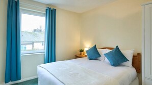 A hotel room with a large bed, blue curtains, a bedside lamp, and a view of a building outside. - Host Stay Trinity Way (Keswick)