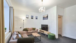 A modern living room with a sectional sofa, a coffee table, a TV, and a chandelier. - Host Stay Queen Street Apartment (Morpeth)