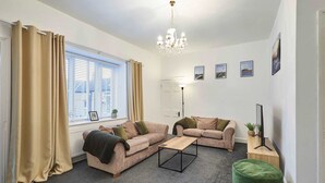 A living room with two sofas, a coffee table, a green ottoman, a chandelier, and a window with curtains. - Host Stay Queen Street Apartment (Morpeth)