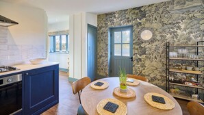 A dining area with a round table, chairs, and placemats. There is a kitchen area with cabinets and a stove. A door with a floral wallpapered wall is visible in the background. - Host Stay The Lodge (Whitby)