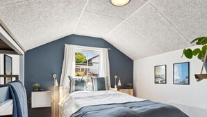 A bedroom with a sloped ceiling, a bed with a dark blue bedspread, a window with curtains, bedside lamps, and framed pictures on the wall. - "Mari" - 150m from the sea by Interhome (Tranekær)