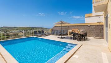 A swimming pool with lounge chairs and a table, surrounded by a white fence, with a view of the landscape.