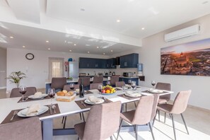Un'area pranzo moderna con un tavolo grande apparecchiato per un pasto, una cucina sullo sfondo e un dipinto con una veduta cittadina sulla parete.