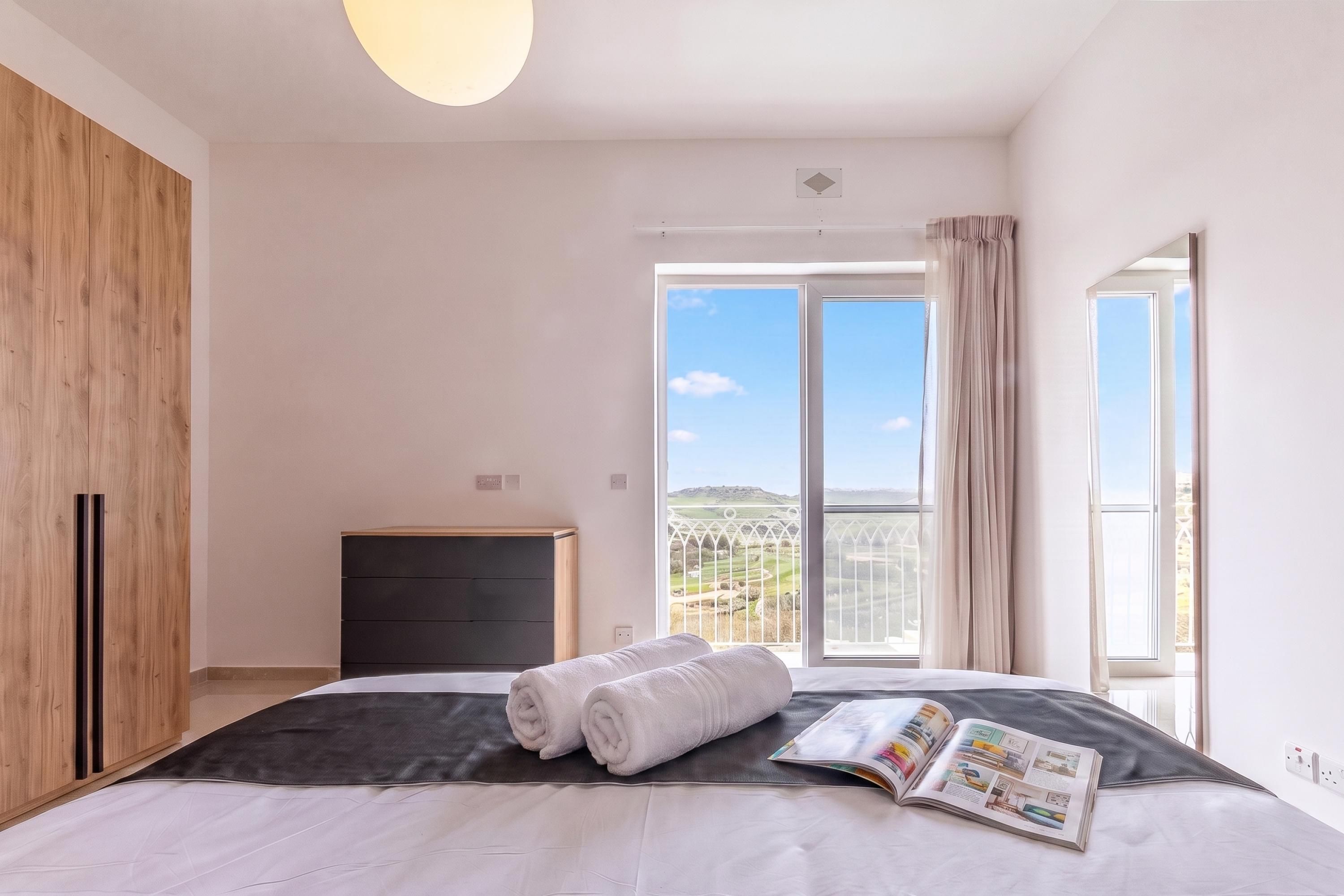 A bedroom with a large bed, a wooden wardrobe, a dark dresser, and a view of the countryside through a large window.