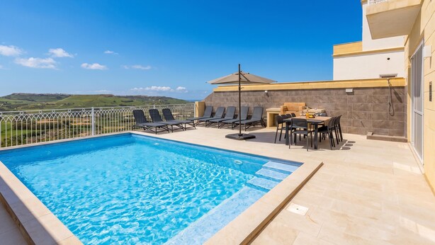 A rooftop pool with clear blue water, surrounded by lounge chairs and a table with chairs, set against a backdrop of a clear sky and distant hills. - Magnifica Gozitan Villa Pool And Jacuzzi (Xaghra)