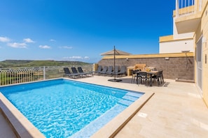 A rooftop pool with clear blue water, surrounded by lounge chairs and a table with chairs, set against a backdrop of a clear sky and distant hills. - Magnifica Gozitan Villa Pool And Jacuzzi (Xaghra)