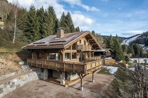 A wooden cabin with a balcony, set on a stone foundation, surrounded by trees and a snowy landscape.