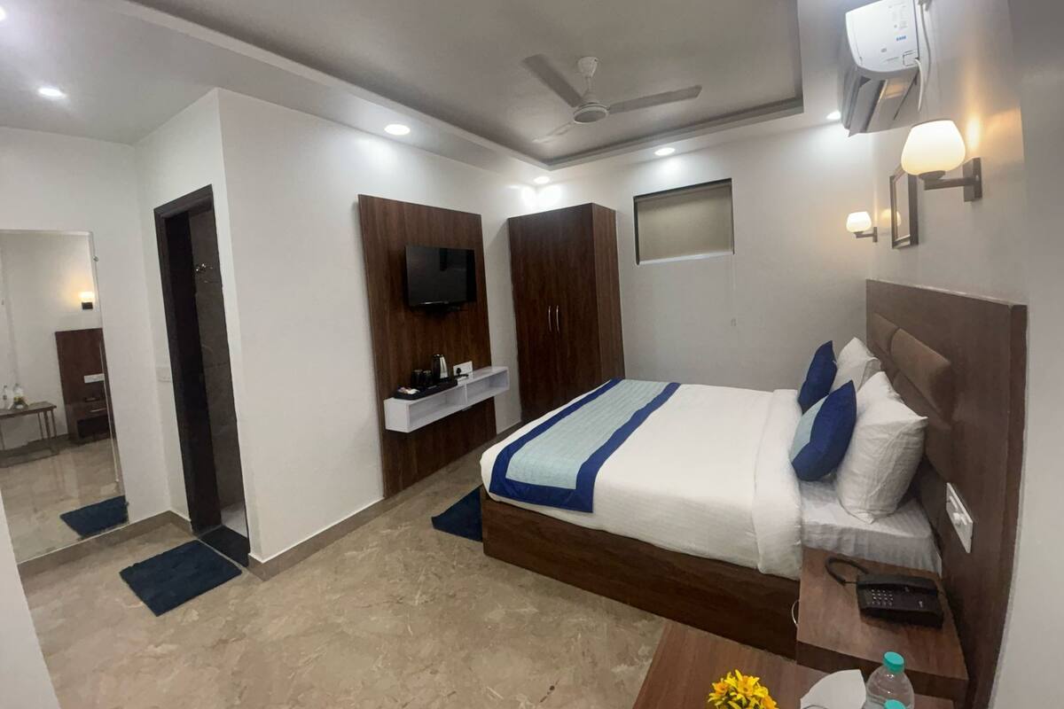 A hotel room with a bed, bedside table, TV, wardrobe, and a small desk with a lamp.