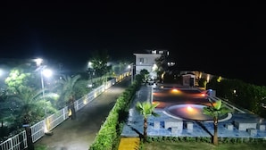 A nighttime view of a well-lit pathway leading to a building with a circular driveway and a small garden area with palm trees. - The Nature View Resort (Kaladhungi)