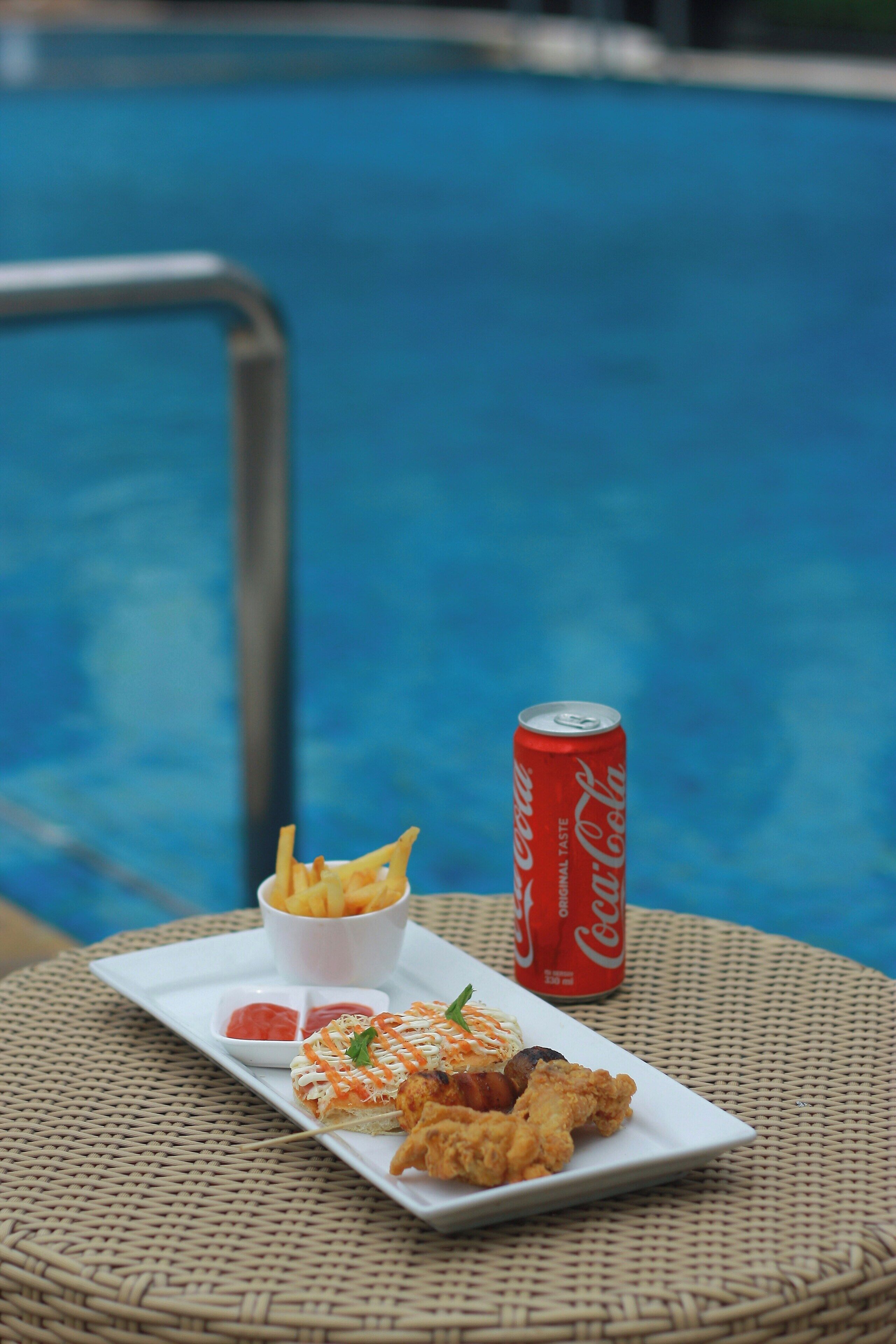 A plate of fried chicken and fries with ketchup and a drink on a wicker table by a pool.