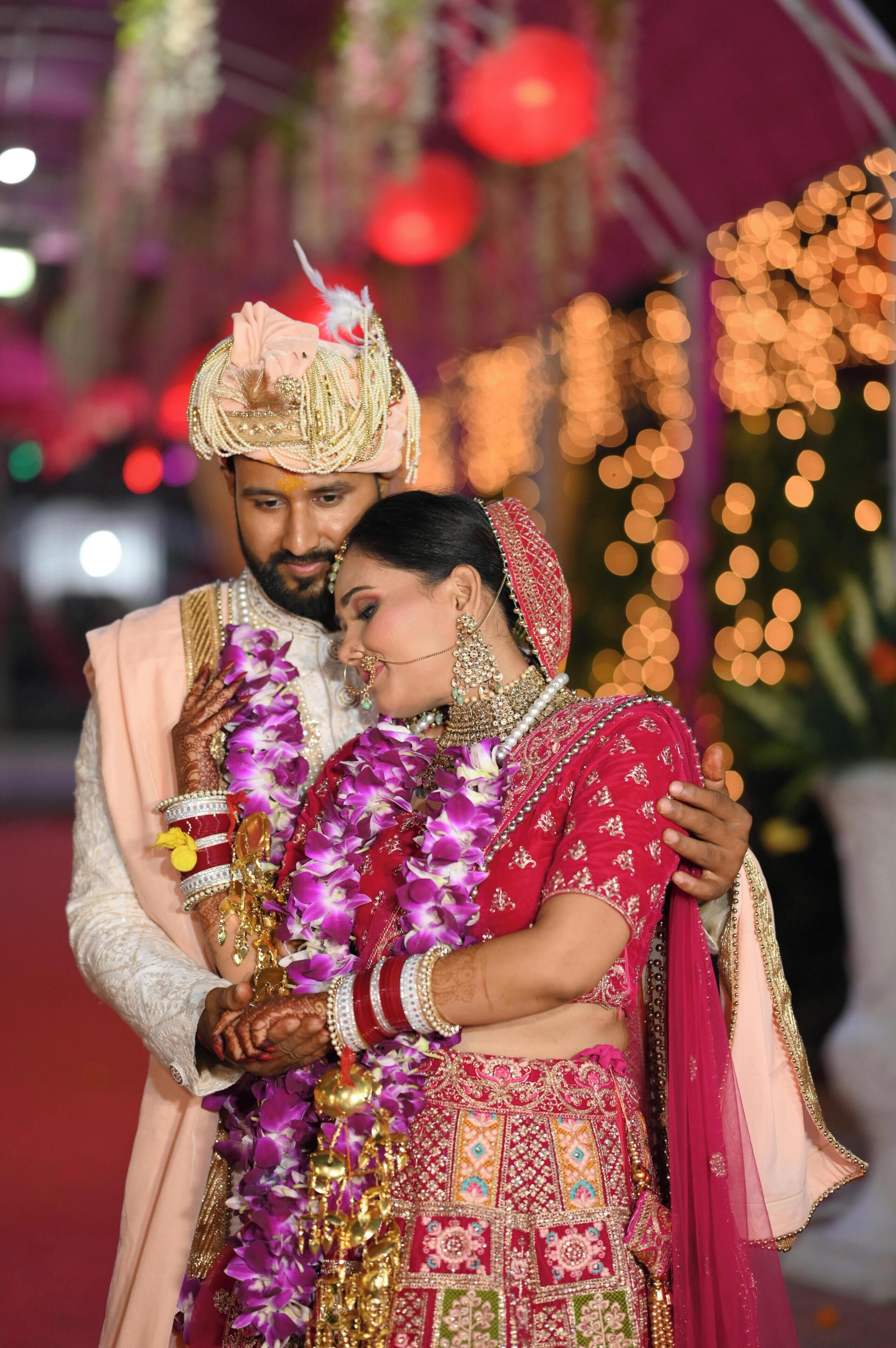 A couple in traditional wedding attire, with the groom in a cream sherwani and the bride in a red lehenga, both adorned with jewelry and garlands.