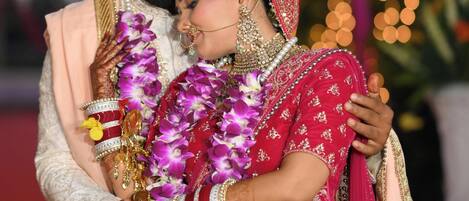 A couple in traditional wedding attire, with the groom in a cream sherwani and the bride in a red lehenga, both adorned with jewelry and garlands.