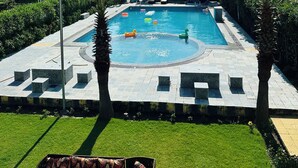 A swimming pool with a clear blue sky, surrounded by palm trees and a grassy area. - The Nature View Resort (Kaladhungi)