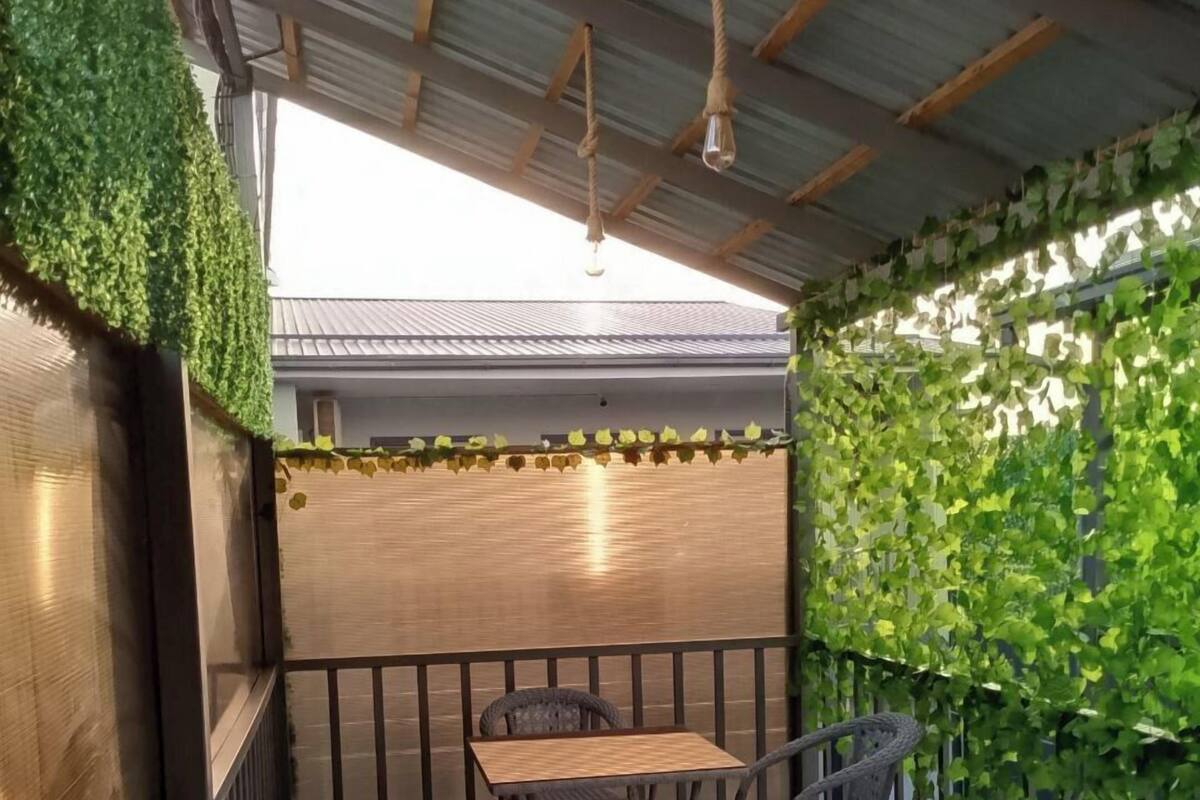 A balcony with a wooden table and two wicker chairs, surrounded by a metal railing and covered with green foliage.