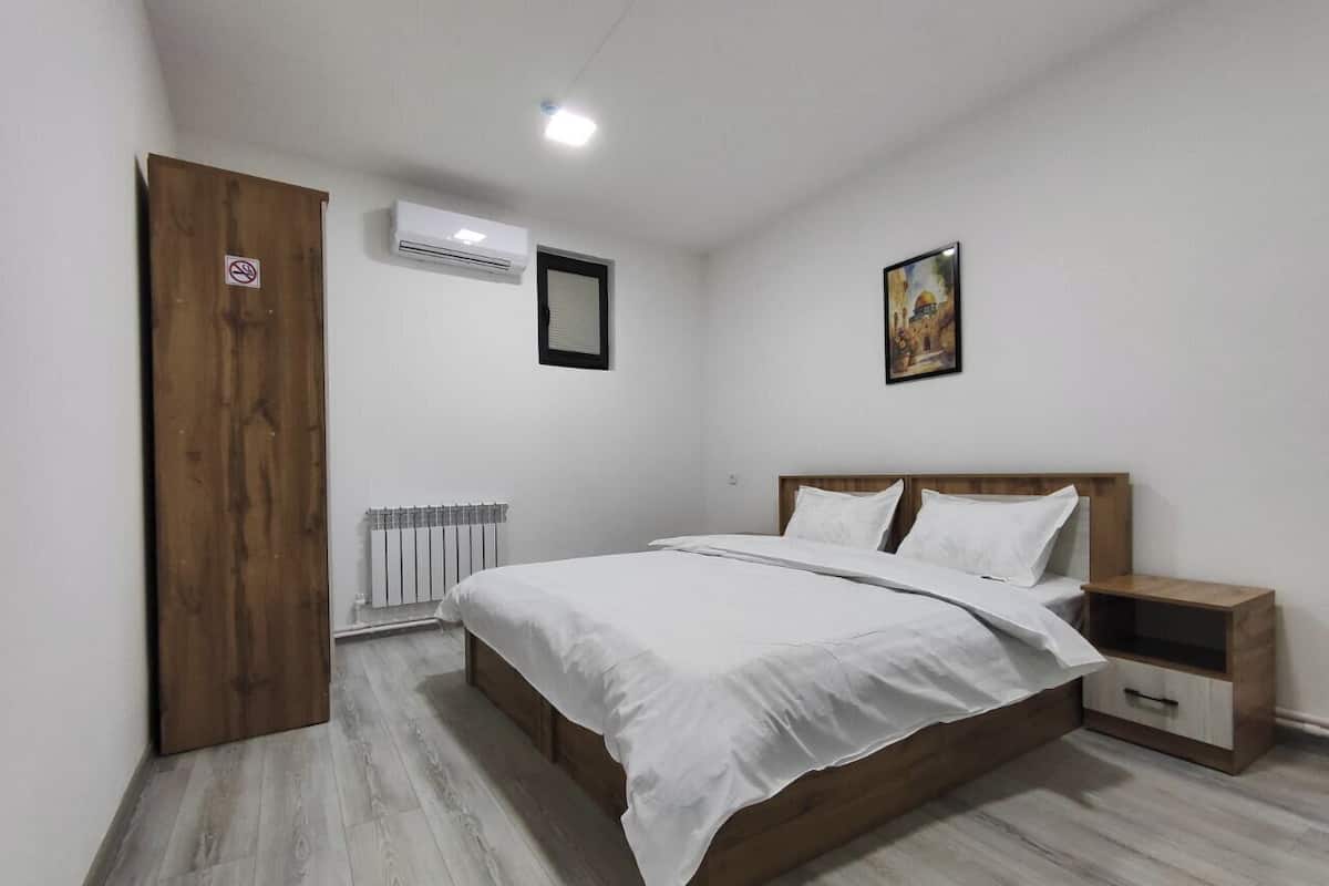 A bedroom with a wooden bed, white bedding, a nightstand, a radiator, and a door with a no-smoking sign.