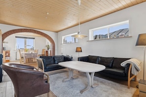 Una sala de estar moderna con techo de madera, un sofá de cuero negro, una mesa de centro blanca y un área de comedor al fondo.