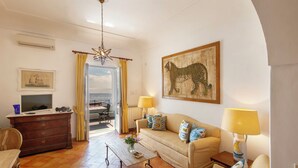 Ein Wohnzimmer mit Sofa, Couchtisch und Fernseher auf einer Kommode. An der Wand hängt ein Tigergemälde und ein Lampenschirm steht auf einem Beistelltisch.