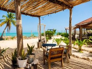 Eine Terrasse am Strand mit einem Holztisch und einer Bank, umgeben von Blumentöpfen und einer Reetdachkonstruktion.