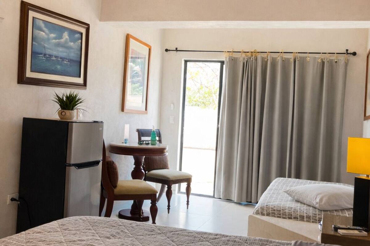 Une chambre d’hôtel comprenant un lit, un bureau, une chaise, une petite table, un climatiseur et une fenêtre avec des rideaux.
