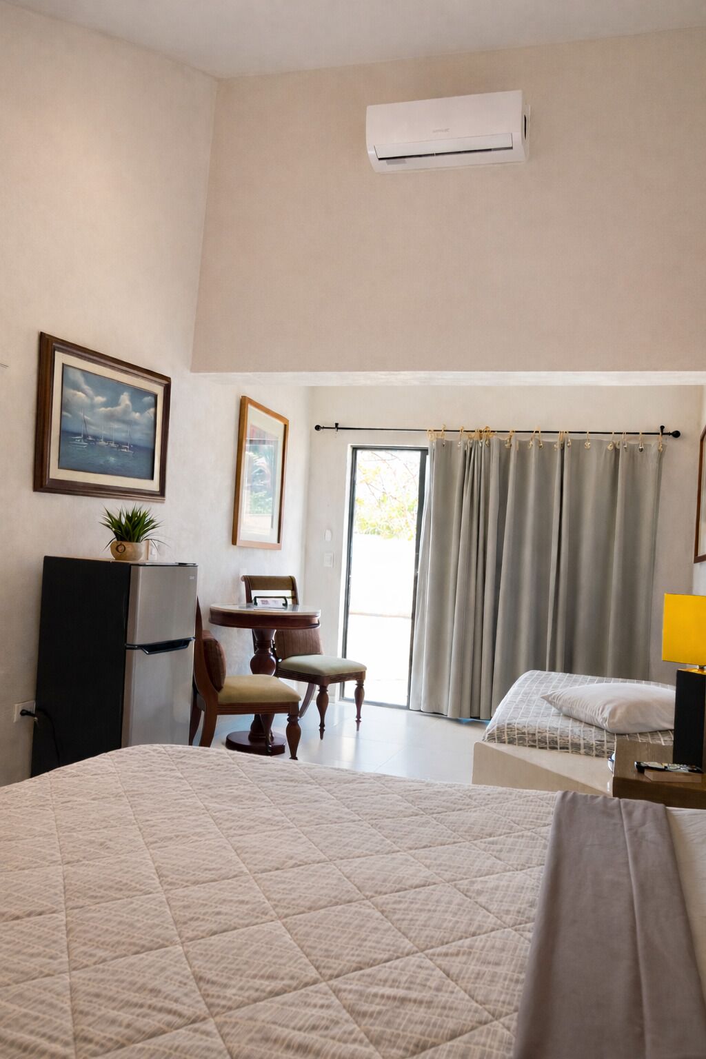 Habitación de hotel con cama, escritorio, silla, refrigerador, ventana con cortinas y aire acondicionado.