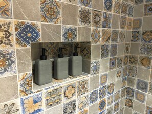 A bathroom with a tiled wall featuring a variety of patterns and a shelf with three gray dispensers. - Bremar Collection Boutique Hotel (Celestún)