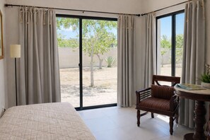 A bedroom with a bed, a chair, a table, and a view of a courtyard with trees. - Bremar Collection Boutique Hotel (Celestún)