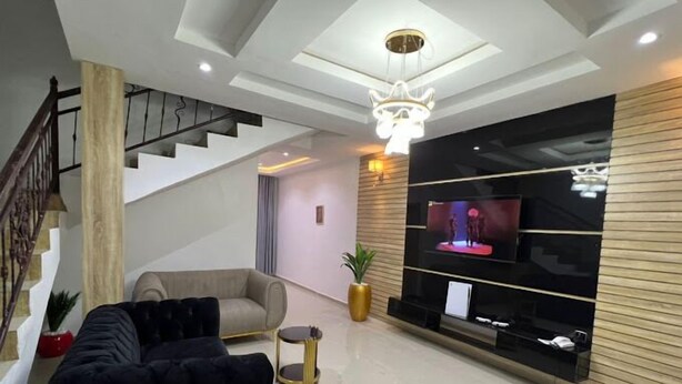 A modern living room with a black sofa, a staircase, a chandelier, and a large flat-screen TV mounted on a wooden wall. - AZ KINGS HOTEL AND APARTMENT (Benin City)
