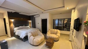 A modern bedroom with a large bed, two armchairs, a small wooden table, and a wall-mounted TV. - AZ KINGS HOTEL AND APARTMENT (Benin City)