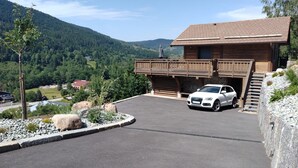 A modern building with a car parked in front, surrounded by a paved area and a mountainous landscape. - Chalet Le Mirador - Sauna - Hammam (Ventron)